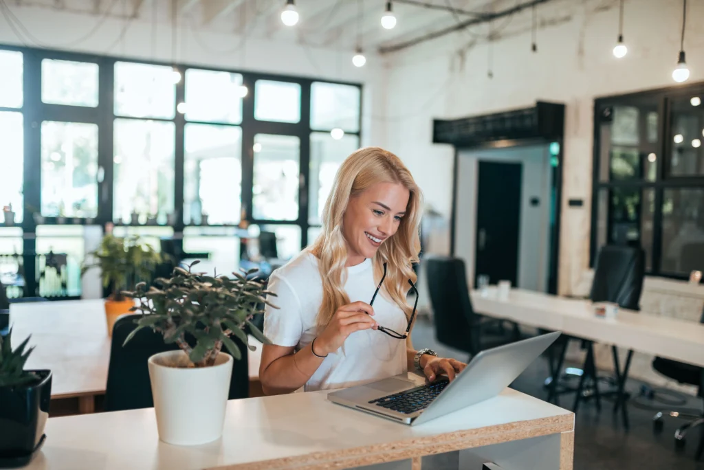 Femme travaillant sur un ordinateur portable dans un bureau lumineux, au téléphone, avec des documents sur le bureau.