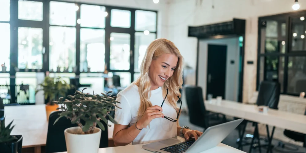 Femme travaillant sur un ordinateur portable dans un bureau lumineux, au téléphone, avec des documents sur le bureau.