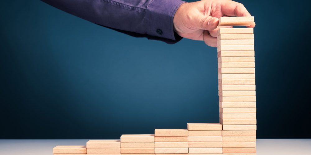 Main empilant des blocs de bois en escalier sur fond bleu, symbolisant la croissance et la finance durable pilotées par l’expert-comptable et le CAC.